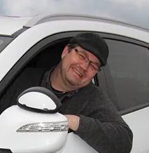 Smilende mand med kasket og briller, der læner sig ud ad vinduet på en hvid bil